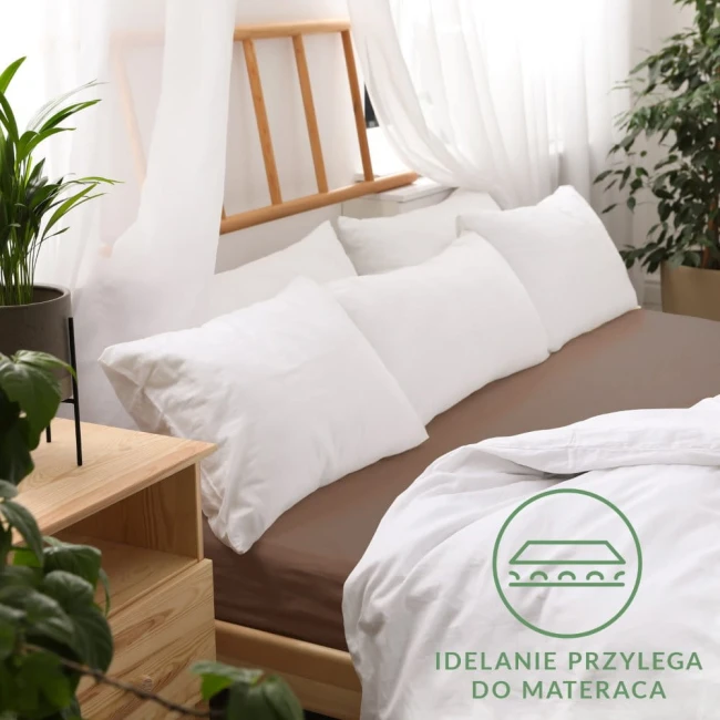 Prześcieradło jersey z gumką 140x200 w kolorze czekoladowym, leżące na łóżku. Na prześcieradle widoczne białe poduszki i część białej pościeli. Tło jasne z drewnianą ramą łóżka i roślinnością. Widoczny napis Idealnie przylega do materaca.