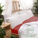 Prześcieradło jersey z gumką 180x200 w kolorze czerwonym, rozłożone na łóżku z białymi poduszkami. Widoczne gładkie wykończenie oraz logo z napisem IDEALNIE PRZYLEGA DO MATERACA w dolnej części zdjęcia. Tło stanowi jasne wnętrze z roślinami.