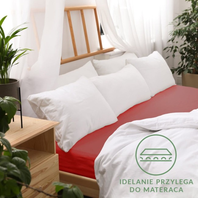 Prześcieradło jersey z gumką 180x200 w kolorze czerwonym, rozłożone na łóżku z białymi poduszkami. Widoczne gładkie wykończenie oraz logo z napisem IDEALNIE PRZYLEGA DO MATERACA w dolnej części zdjęcia. Tło stanowi jasne wnętrze z roślinami.