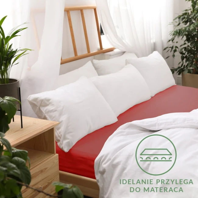 Prześcieradło jersey z gumką 140x200 w kolorze czerwonym, rozłożone na łóżku, z wyraźnym podbiciem krawędzi. W tle białe poduszki i firany, stół nocny z rośliną. Widoczny napis IDEALNIE PRZYLEGA DO MATERACA na białym tle.