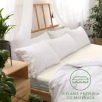 Prześcieradło jersey z gumką w kolorze ecru, rozmiar 200x220 cm, leżące na łóżku z poduszkami. Tło z drewnianym łóżkiem i roślinami. Widoczna tekstura materiału, idealnie przylegająca do materaca, z logo i napisem w dolnym rogu.