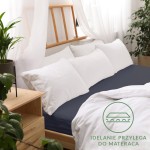 Prześcieradło jersey z gumką w kolorze ciemnego granatu, rozmiar 180x200 cm, leży na łóżku z białą pościelą oraz poduszkami. Otoczenie to jasne wnętrze z drewnianą ramą łóżka i roślinnością. Na materiale widoczna faktura dzianiny.