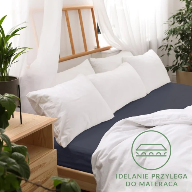 Prześcieradło jersey z gumką w kolorze ciemnego granatu, rozmiar 180x200 cm, leży na łóżku z białą pościelą oraz poduszkami. Otoczenie to jasne wnętrze z drewnianą ramą łóżka i roślinnością. Na materiale widoczna faktura dzianiny.