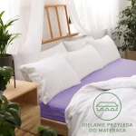 Prześcieradło jersey z gumką w kolorze jasnego fioletu leży na łóżku, otoczone białymi poduszkami. Widoczne jest odpowiednie dopasowanie do materaca. Tło to drewniane łóżko i rośliny, co nadaje aranżacji przytulny charakter.