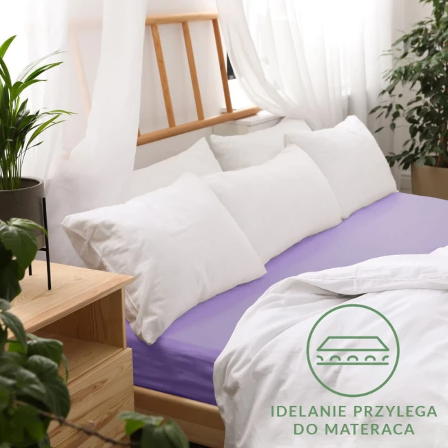 Prześcieradło jersey z gumką w kolorze jasnego fioletu leży na łóżku, otoczone białymi poduszkami. Widoczne jest odpowiednie dopasowanie do materaca. Tło to drewniane łóżko i rośliny, co nadaje aranżacji przytulny charakter.