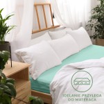 Prześcieradło jersey z gumką 160x200 w jasnym turkusie, rozłożone na łóżku z białymi poduszkami. Otoczenie z drewnianą ramą wezgłowia i roślinami w doniczkach. Widoczne logo z hasłem „Idealnie przylega do materaca”.