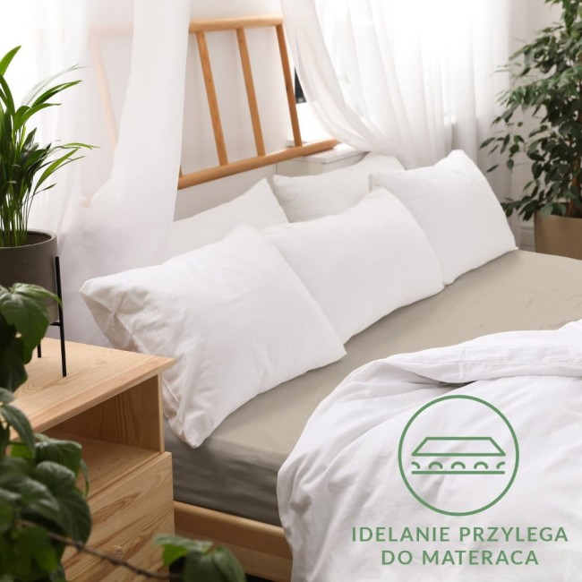 Prześcieradło jersey z gumką 140x200 w kolorze kakao, leżące na łóżku z poduszkami w białych poszewkach. Widoczny jest detal gumki zapewniającej stabilność. Tło w jasnych barwach, na stole rośliny doniczkowe oraz drewniane akcesoria.