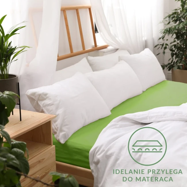 Prześcieradło jersey z gumką w kolorze kaktusowym, rozłożone na łóżku z poduszkami o białych poszewkach. Kąt ujęcia z boku, na tle jasnego wnętrza z rośliną i drewnianym meblem. Widoczne szczegóły tkaniny oraz nadruk informacyjny o właściwościach.