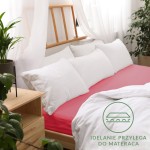 Prześcieradło jersey z gumką w kolorze koralowym, leżące na łóżku, oplata materac, widoczne białe poduszki i narzuta. Tło z drewnianą ramą łóżka oraz roślinami, logo z napisem „Idealnie przylega do materaca” w dolnym rogu.