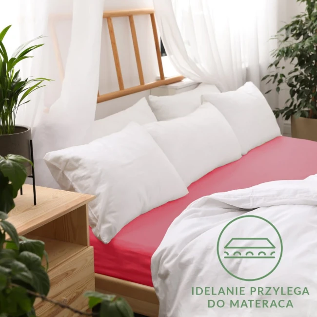 Prześcieradło jersey z gumką w kolorze koralowym, leżące na łóżku, oplata materac, widoczne białe poduszki i narzuta. Tło z drewnianą ramą łóżka oraz roślinami, logo z napisem „Idealnie przylega do materaca” w dolnym rogu.