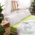Prześcieradło jersey z gumką 200x220 w kolorze limonkowym, rozłożone na łóżku z białą pościelą i poduszkami. Widoczny jest naturalny materiał i dopasowanie do materaca. Tło stanowi jasne wnętrze z roślinnością i drewnianymi meblami.