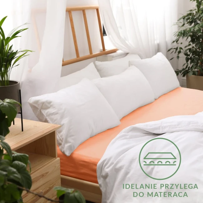 Prześcieradło jersey z gumką w kolorze łososiowym, rozmiar 140x200, leżące na łóżku z białymi poduszkami. Tło jasne z roślinami doniczkowymi i drewnianym meblem. Logo w dolnym rogu: Idealnie przylega do materaca.
