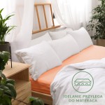 Prześcieradło jersey z gumką w kolorze łososiowym, o wymiarach 200x220 cm, leży na łóżku, podparte na białych poduszkach.