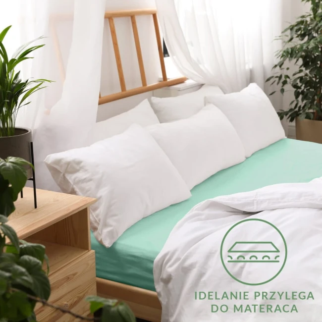 Prześcieradło jersey z gumką w kolorze morskim (80x160 cm) leży na łóżku, otoczone białymi poduszkami i kołdrą. Widoczne gładkie wykończenie oraz elastyczna gumka zapewniająca idealne dopasowanie. Tło z naturalnymi dekoracjami roślinnymi.