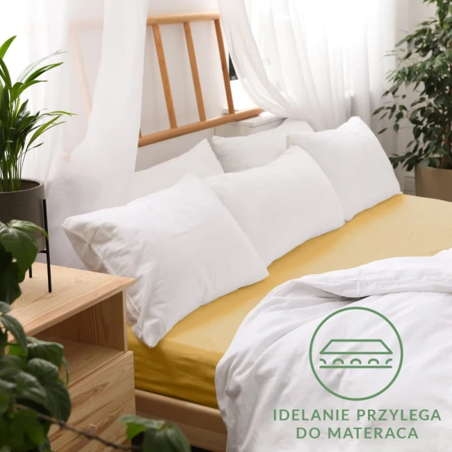 Prześcieradło jersey 180x200 w kolorze musztardowym, rozłożone na łóżku z białymi poduszkami. Widoczna gumka, która napina materiał. Tło jasne, z naturalnym światłem i roślinnością. Obok widoczny drewniany stelaż i komoda.