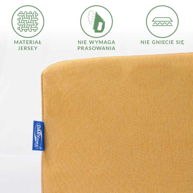 Prześcieradło jersey z gumką w kolorze musztardowym, leżące na białym tle. Zbliżenie na fakturę materiału, z widoczną metką w niebieskim kolorze. Wskazania dotyczące braku potrzeby prasowania oraz odporności na gniecenie.