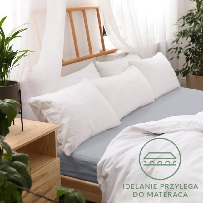 Prześcieradło jersey z gumką 90x200 w kolorze popielatym, rozłożone na łóżku. Widzoczne poduszki w białych poszewkach i lekka narzuta.