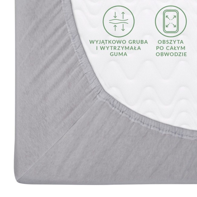 Prześcieradło jersey z gumką w kolorze popielatym, rozmiar 160x200 cm. Ujęcie z góry, ukazujące grube wykończenie gumą i fakturę materiału. Na powierzchni widoczne grafiki informujące o wytrzymałej gumie i obszytowaniu po obwodzie.