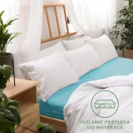 Prześcieradło jersey z gumką 200x220 w kolorze turkusowym, rozłożone na materacu, z białymi poduszkami w tle. Tło jasne, z roślinnością i drewnianym meblem. Widoczne logo w dolnej części zdjęcia, podkreślające doskonałe przyleganie do materaca.