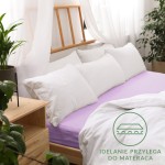 Prześcieradło jersey z gumką w kolorze wrzosowym, rozmiar 200x220 cm, ułożone na materacu z białymi, puszystymi poduszkami. Widoczny nadruk Idealnie przylega do materaca. Tło to jasne wnętrze z drewnianym łóżkiem i roślinami, na prostym tle.