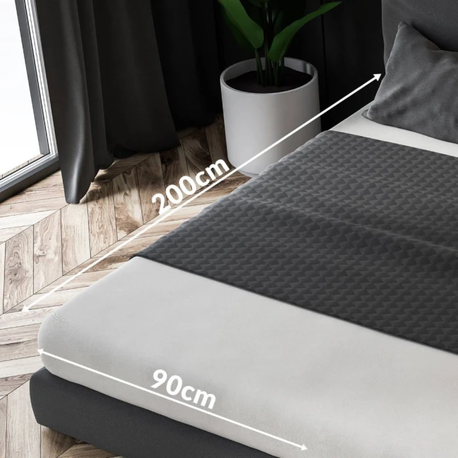 Prześcieradło frotte z gumką w kolorze jasnego beżu o wymiarach 90x200 cm, rozłożone na materacu. Na zdjęciu widoczny jest zbliżenie na fakturę materiału oraz oznaczenia wymiarów. W tle naturalne wnętrze z roślinnością i oknem.