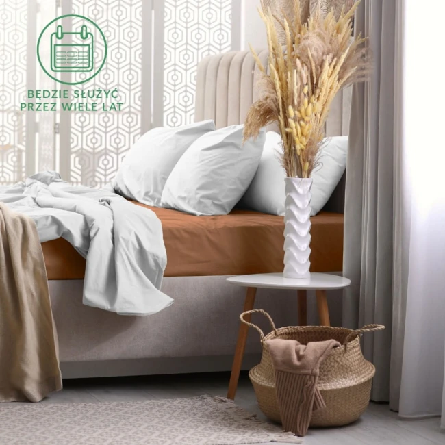 Prześcieradło frotte z gumką 120x200 w kolorze czekoladowym leży na materacu łóżka. Widoczne są poduszki oraz narzuta w jasnym kolorze. Tło to jasna, zdobiona ściana oraz stół z kwiatami w wazonie i koszem.