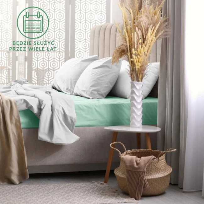 Prześcieradło frotte z gumką 120x200 w kolorze morskim, rozłożone na łóżku z szarymi poduszkami. Otoczenie to elegancka sypialnia z jasnymi zasłonami, białą podstawką i wazonem z wyschniętymi kwiatami. Tło z delikatnym wzorem.