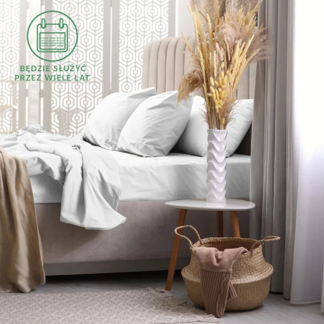 Prześcieradło frotte z gumką 140x200 w kolorze białym, leżące na łóżku z poduszkami o jasnym odcieniu. W tle dekoracyjna ścianka i stół z wazonem oraz koszem, tworzące przytulną aranżację sypialni.