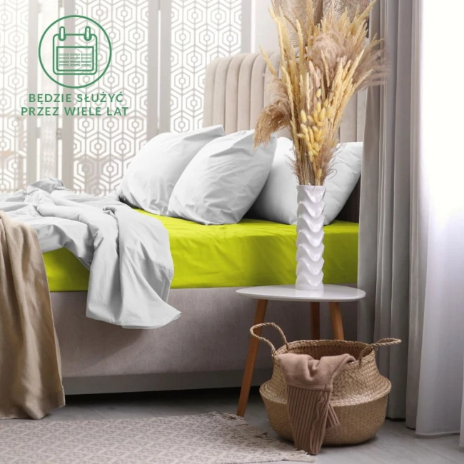 Prześcieradło frotte z gumką 140x200 w kolorze limonkowym, leżące na łóżku. Pościel biała w tle, na stoliku biały wazon z dekoracyjnymi trawami. Widoczne miękkie tekstury materiałów, estetyka nowoczesnej sypialni.