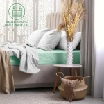 Prześcieradło frotte z gumką 140x200 w kolorze morskim, rozłożone na łóżku z białymi poszewkami, widok z boku. W tle wazon z suszonymi roślinami na stoliczku oraz kosz z dodatkowymi akcesoriami. Proste tło z jasnymi zasłonami.