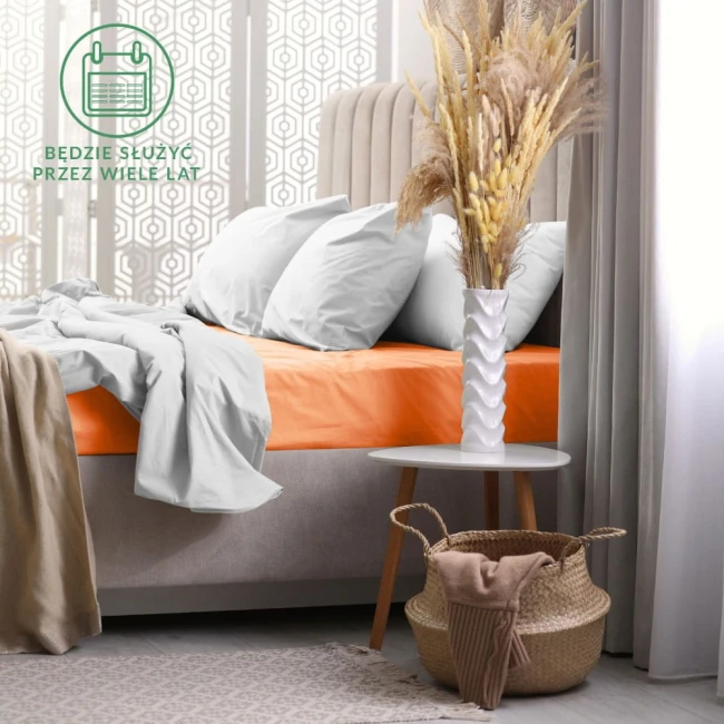 Prześcieradło frotte z gumką 140x200 w kolorze łososiowym, leżące na łóżku. Widoczne gładkie, elastyczne tkaniny, otoczone jasnymi poduszkami. Dopełnieniem aranżacji jest biała waza z suszonymi kwiatami na stoliku i kosz z materiałem obok.
