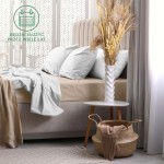 Prześcieradło frotte z gumką 160x200 w kolorze beżowym. Leży na łóżku, częściowo zwinięte, z widocznymi poduszkami w jasnym odcieniu. W tle dekoracje i stół z wazonem oraz suszonymi kwiatami. Tło jasne i przyjemne, podłoga drewniana.