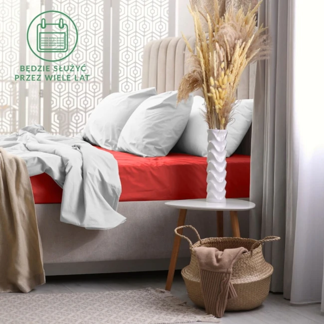 Prześcieradło frotte z gumką 160x200 w kolorze czerwonym, rozłożone na łóżku z jasną pościelą. Kąt ujęcia frontalny, podkreślający fakturę materiału. W tle białe zasłony, stół nocny z wazonem i suszonymi kwiatami. Widzoczny kosz z materiałem obok.