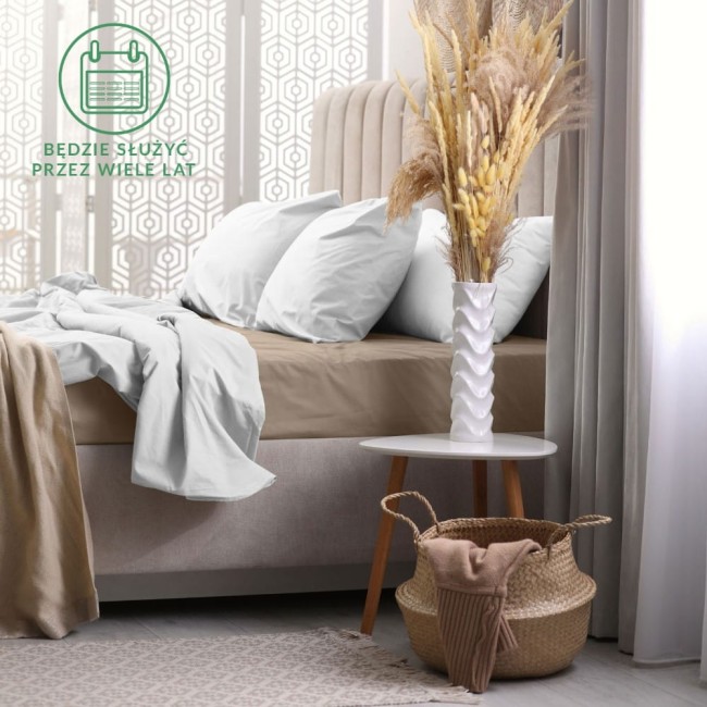 Prześcieradło frotte z gumką w kolorze kakao, rozłożone na łóżku z jasnymi poduszkami. Ujęcie frontalne, w tle widoczna dekoracyjna zasłona i stół z dekoracją. Produkt jest wykonany z miękkiego materiału, idealny do łóżka o wymiarach 160x200.