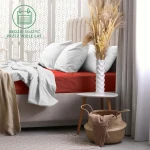 Prześcieradło frotte z gumką w kolorze bordo rozłożone na łóżku z jasnym, gładkim materacem. W tle widoczne poduszki w kolorze białym oraz dekoracyjny wazon z suszonymi roślinami na białym stoliku. Tło utrzymane w jasnych tonach.