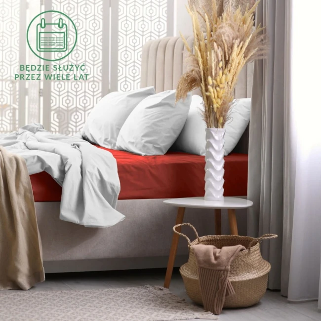 Prześcieradło frotte z gumką w kolorze bordo rozłożone na łóżku z jasnym, gładkim materacem. W tle widoczne poduszki w kolorze białym oraz dekoracyjny wazon z suszonymi roślinami na białym stoliku. Tło utrzymane w jasnych tonach.