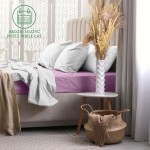 Prześcieradło frotte z gumką 180x200 w jasnym fiolecie leży na łóżku, obok poduszki w białych poszewkach. Ułożenie jest rozłożone, na tle beżowego zagłówka oraz kremowych tkanin. Widoczny dekoracyjny wazon z trzciną i stolik w jasnym kolorze.