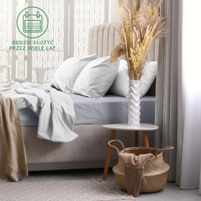 Prześcieradło frotte z gumką 180x200 w kolorze popiel szaro-niebieskim, złożone na łóżku. Na zdjęciu widoczne również poduszki oraz naturalne dekoracje, takie jak stół z białym wazonem i suszonymi kwiatami. Tło jasne, stylowe wnętrze.