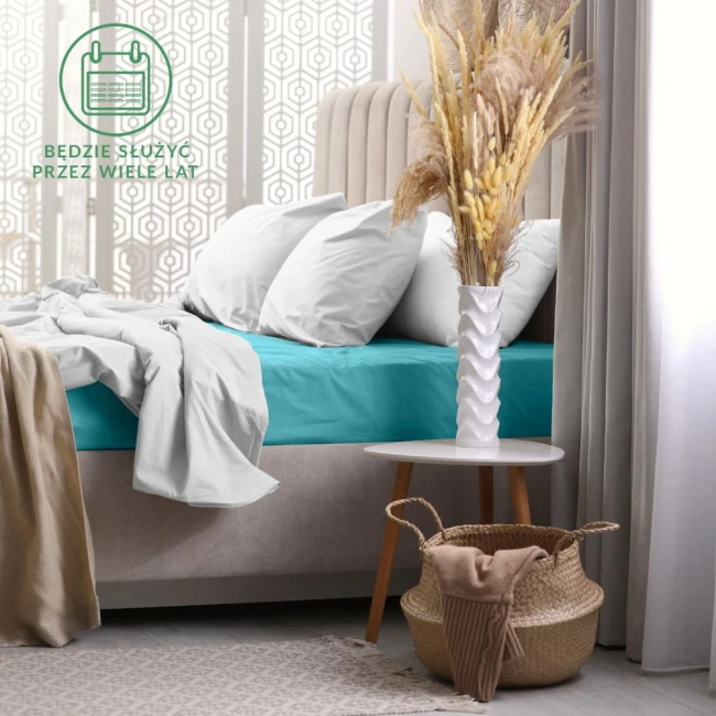 Prześcieradło frotte z gumką w kolorze turkusowym, o wymiarach 180x200 cm, rozłożone na łóżku z białymi poduszkami. W tle widoczny stół z dekoracyjną wazonem i suszonymi roślinami, na jasnym tle.