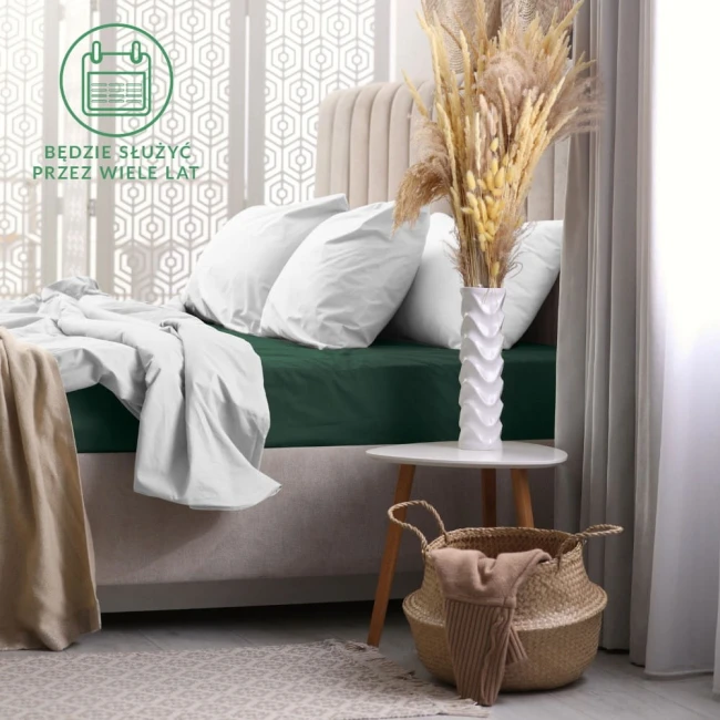 Prześcieradło frotte z gumką w kolorze zieleni butelkowej o wymiarach 180x200 cm, rozłożone na łóżku. W tle widoczne poduszki w białym poszyciu oraz dekoracyjne elementy, takie jak wazon z suszonymi kwiatami i kosz.
