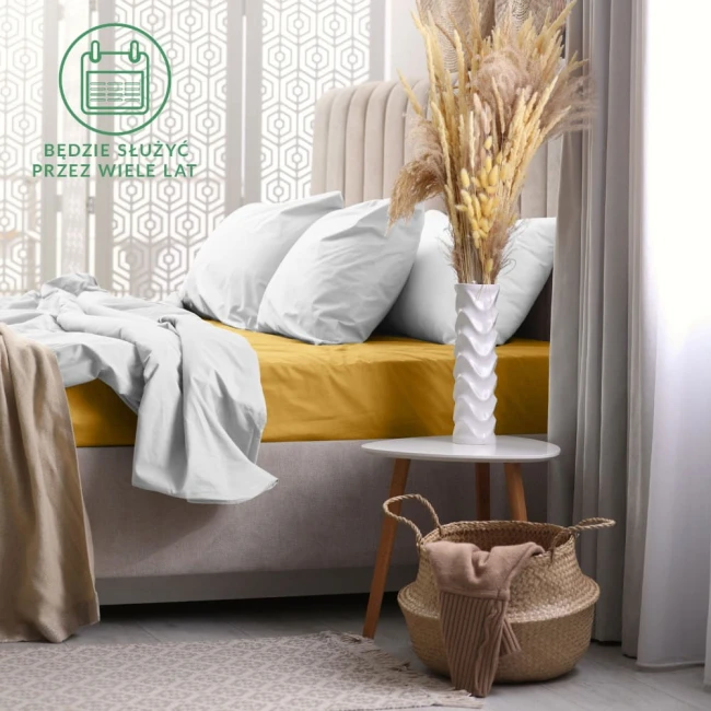 Prześcieradło frotte z gumką w kolorze musztardowym, rozłożone na łóżku z białymi poduszkami. W tle widoczny stylowy zagłówek oraz stół nocny z wazonem i dekoracją. Całość w jasnym, nowoczesnym wnętrzu z drewnianą podłogą.