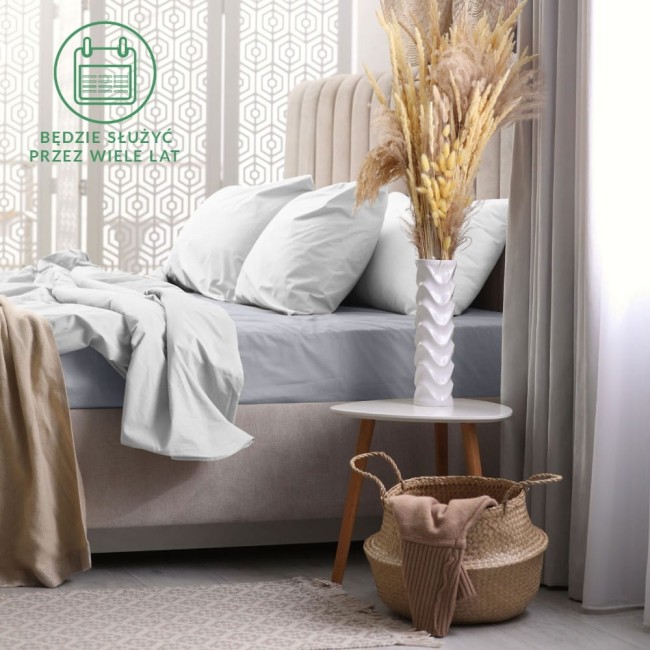 Prześcieradło frotte z gumką w kolorze popielatym o wymiarach 200x220 cm, leżące na łóżku z jasną pościelą. W tle widoczna dekoracyjna waza z suszonymi kwiatami oraz kosz z tekstyliami. Stonowane, nowoczesne wnętrze.