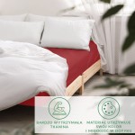 Prześcieradło satynowe z gumką w kolorze czerwonym, rozmiar 90x200 cm, leżące na łóżku. Widać białą pościel i poduszki, a także drewnianą konstrukcję łóżka. Tło jasne, z oknem i zasłonami.