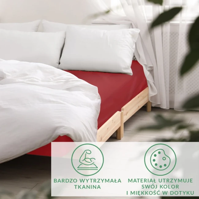 Prześcieradło satynowe z gumką w kolorze czerwonym, rozmiar 90x200 cm, leżące na łóżku. Widać białą pościel i poduszki, a także drewnianą konstrukcję łóżka. Tło jasne, z oknem i zasłonami.