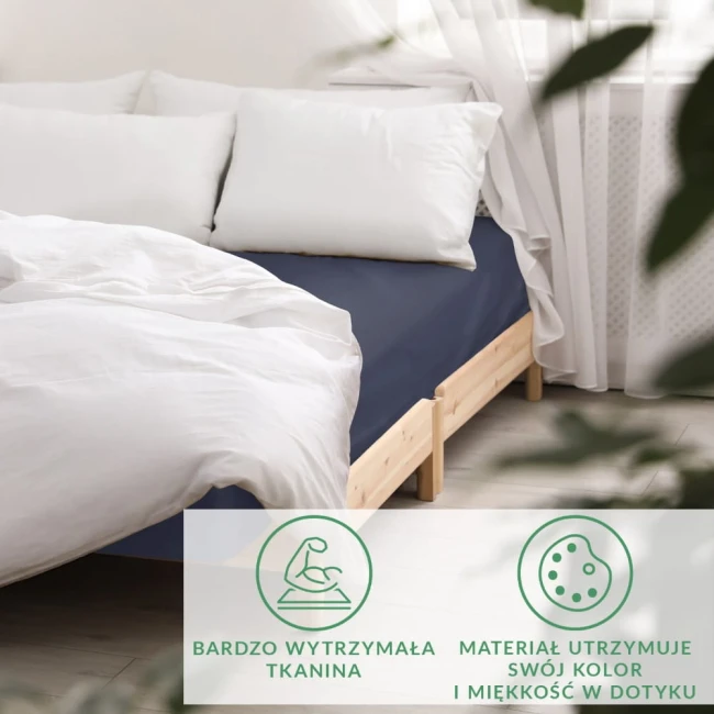 Prześcieradło satynowe z gumką w ciemnym granacie, leżące na łóżku z drewnianą ramą. Widoczne poduszki w białych poszewkach. Tło jasne, z firankami i roślinami dekoracyjnymi, podkreślające elegancki wygląd aranżacji sypialni.