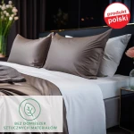 Prześcieradło satynowe z gumką 140x200 w kolorze czarnym, układ leżący. Widoczne poduszki w odcieniu brązowym oraz białym. Na tle eleganckiej sypialni z ciemnymi zasłonami. Wyraźny nadruk Bez domieszek sztucznych materiałów w dolnej części zdjęcia.