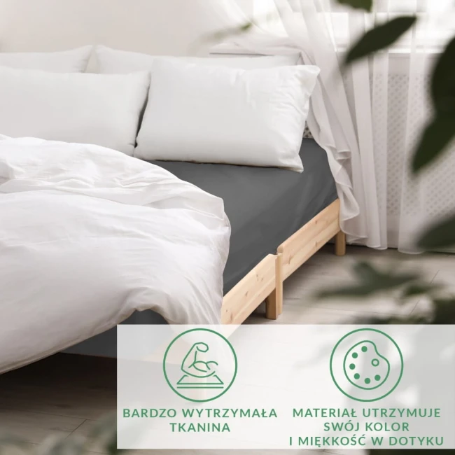 Szare prześcieradło satynowe z gumką rozłożone na łóżku, w otoczeniu białych poduszek i jasnej pościeli. Ujęcie frontalne, prezentujące fakturę materiału.