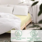 Prześcieradło satynowe z gumką 180x200 w kolorze kremowym, rozłożone na łóżku z białą pościelą. Wykonane z trwałej tkaniny, posiada gładką, miękką fakturę. Widoczne w tle naturalne światło i białe zasłony.