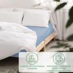 Prześcieradło satynowe z gumką w kolorze błękitnym, rozłożone na łóżku z naturalnego drewna. Widoczna gładka powierzchnia tkaniny, idealnie dopasowane do materaca. W tle białe poduszki i zasłony, na prostym, jasnym tle.