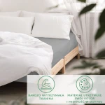 Prześcieradło satynowe z gumką w kolorze popielatym, rozłożone na łóżku o drewnianej ramie, z białą pościelą i poduszkami. W tle zasłony oraz rośliny. Widoczna faktura tkaniny. Idealne do sypialni.